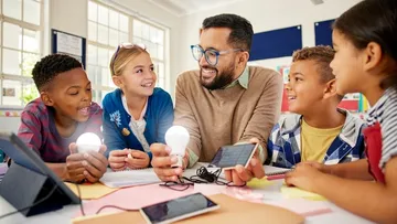 Sieben Personen arbeiten in einem hellen Büro mit Pflanzen und Fenstern. Sie haben Laptops, Notizbücher und Haftnotizen auf den Tischen. Sie tragen bunte Kleidung.