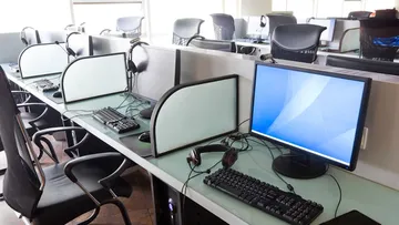 Ein Büroarbeitsplatz mit mehreren Schreibtischen. Jeder Tisch hat einen Stuhl, einen Computerbildschirm, eine Tastatur, eine Maus und Kopfhörer. Die Schreibtische sind durch kleine Wände getrennt. Große Fenster lassen viel Licht herein. Der Boden ist mit dunklen Fliesen bedeckt.