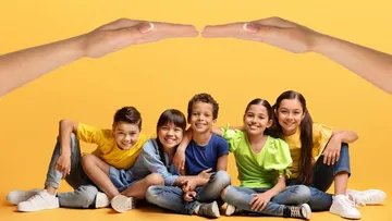 Fünf lachende Kinder sitzen zusammen vor einem gelben Hintergrund, über ihnen halten zwei Hände wie ein schützendes Dach.