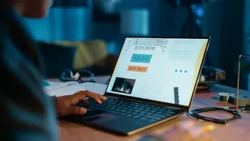 Eine Person arbeitet an einem Laptop in einem dunklen Raum. Der Bildschirm zeigt Formeln und ein Bild. Auf dem Tisch liegen eine Brille, Papiere und ein Gerät mit Kabeln. Im Hintergrund sieht man eine gelbe Couch.