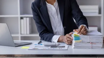 Eine Person ordnet Papierstapel neben einem Laptop.