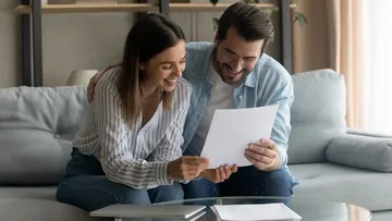 Ein Paar sitzt auf einem Sofa und schaut auf ein Blatt Papier. Die Frau trägt eine gestreifte Bluse und Jeans, der Mann eine blaue Jacke und Jeans. Auf dem Tisch liegen ein Laptop und Papiere. Im Hintergrund sieht man Regale mit Büchern und eine Pflanze.