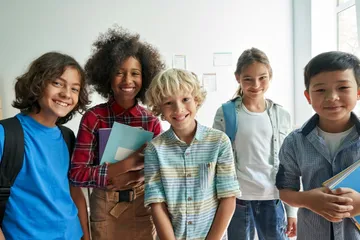 Fünf lachende Kinder stehen im Klassenzimmer mit Notizbüchern und Rucksäcken. Sie tragen bunte Kleidung. Ein Fenster lässt Licht herein.