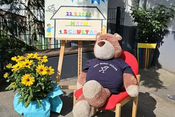 Teddy auf einem Stuhl vor Einschultafel 2024 daneben gelbe Blumen