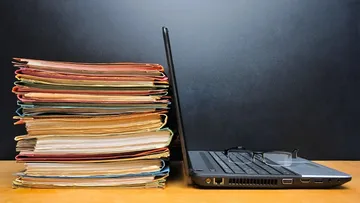 Viele Akten liegen neben einem Laptop mit einer Brille darauf.